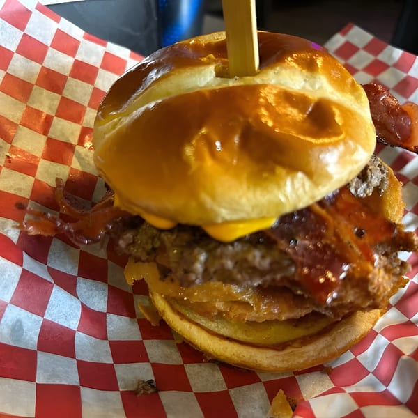A bacon cheeseburger with a wooden skewer, served on red and white checkered paper.