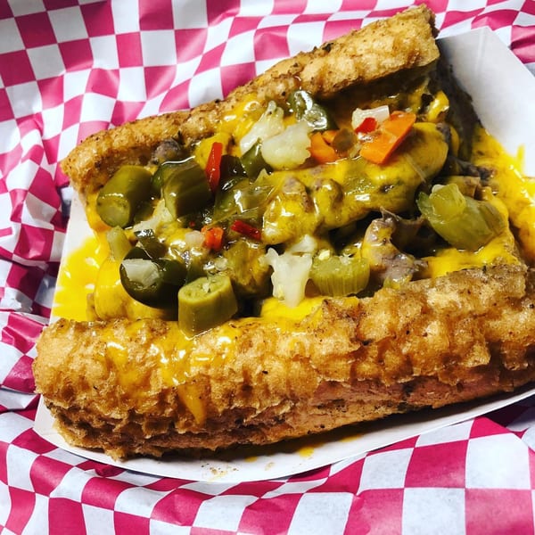 Cheesesteak sandwich with melted cheese, pickled vegetables, and jalapeños in a red and white checkered paper.