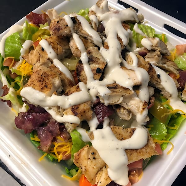 Grilled chicken salad with bacon, cheese, and ranch dressing in a white takeout container.
