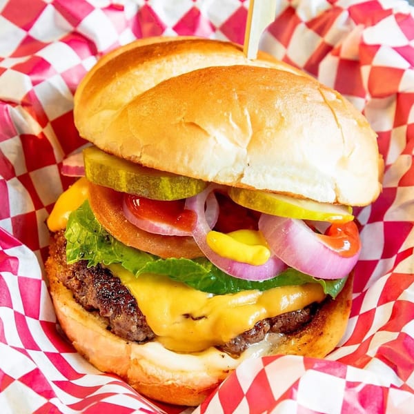 Cheeseburger with lettuce, tomato, onion, pickles, ketchup, and mustard in red and white checkered paper.