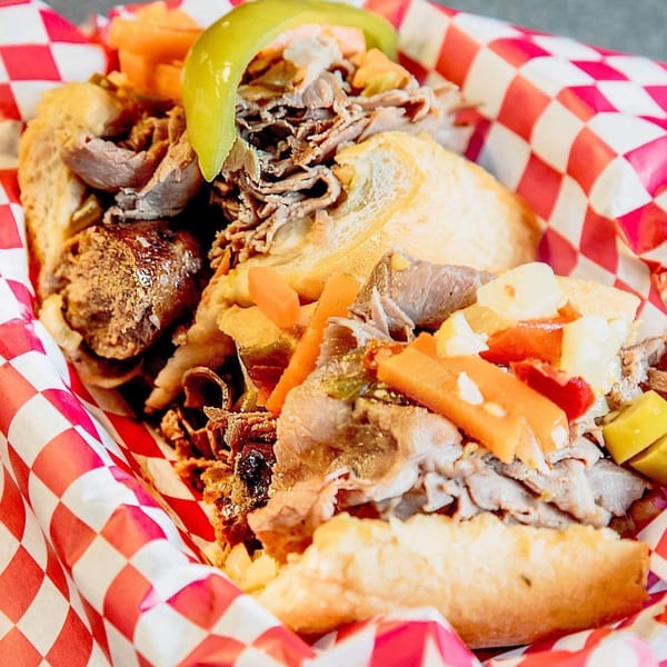 Italian beef sandwich with sausage, giardiniera, and a pepperoncini in a red and white checkered basket.